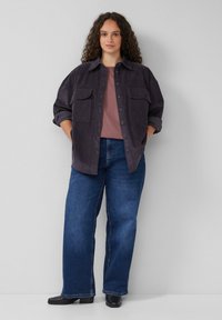 Donkerpaarse corduroy shirt met twee voorzakken, gelaagd over een mauve top, gecombineerd met wijde blauwe jeans en zwarte enkelboots.