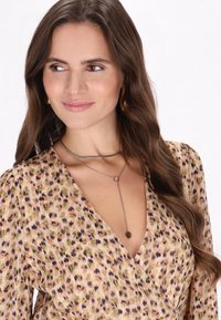 Zilveren gelaagde ketting met een ronde hanger en een ronde, gestructureerde accent; gedragen over een wikkeljurk met een bloemenpatroon in gedempte kleuren.