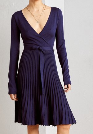 Woman wearing a navy blue long-sleeve pleated wrap dress with a tie belt and layered gold necklaces against a plain background.
