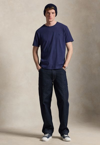 Νέος άνδρας που φοράει μπλε σκούφο, μπλε μπλούζα, σκούρα τζιν, μπλε αθλητικά παπούτσια και ρολόι χειρός, στέκεται με τα χέρια στις τσέπες μπροστά σε απλό φόντο.