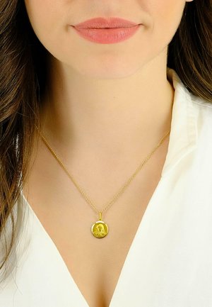 Collana con ciondolo in oro, caratterizzata da un charm circolare con un design inciso, sospesa su una delicata catena contro una camicetta bianca.