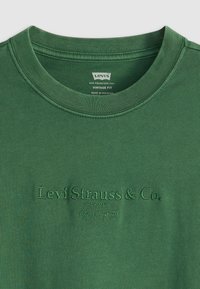 Zelené bavlněné tričko s vintage střihem. Obsahuje žebrovaný kulatý výstřih a vyražené logo, má hladkou texturu a minimalistický design.