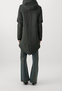 Mörkgrön lång kappa med huva, cirkulär fåll och en dragkedja på baksidan, gjord av ett mjukt tyg. Kombinerad med utsvängda jeans.