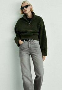 Zeleni flis pulover z visokim ovratnikom in zadrgo, v kombinaciji z visokim pasom svetlo sivimi kavbojkami. Model nosi temne sončne očali.