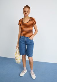 Stripet brun og oransje tettsittende topp, blå denimshorts, hvite sko med nettdesign, holder en heklede veske med gule blomsterdetaljer.