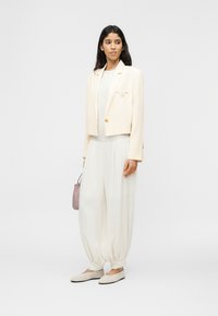 Γυναίκα με λευκό χαλαρό παντελόνι με ρεβέρ στους αστραγάλους, κοντό κρεμ σακάκι, μπεζ μπαλαρίνες και κρατώντας μικρή τσάντα σε μούβ χρώμα, στέκεται μπροστά σε λευκό φόντο.