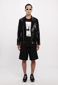 Jachetă de motociclist din piele neagră cu fermoare argintii, tricou grafic alb, shorts negri largi, ochelari de soare albi și adidași negri cu text.