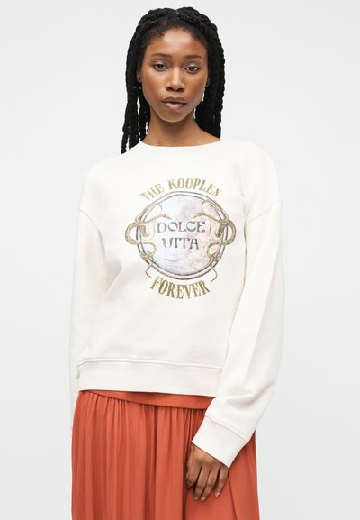 Γυναίκα με μακριά πλεγμένα μαλλιά που φοράει λευκό φούτερ με τύπωμα "The Kooples Dolce Vita Forever" και ροδακινί πλισέ φούστα.