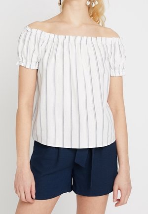 Kvinde iført hvid off-shoulder top med lodrette striber og marineblå højtaljede shorts med frontbindebånd.