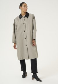 Beige trenčkot s černým límcem, který má velké knoflíky a kapsy s klopami. Model ho nosí přes tmavé kalhoty a špičaté černé kotníkové boty.