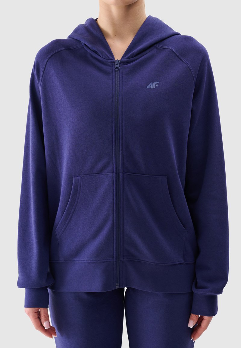 Felpa con zip di colore viola, dotata di cappuccio, realizzata in tessuto morbido, con due tasche frontali e un piccolo logo sul lato sinistro del petto.