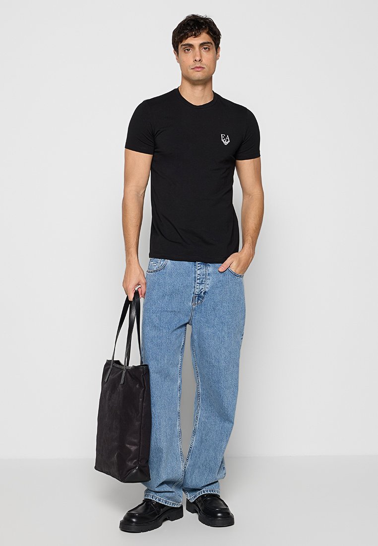 Hombre de pie contra una pared gris con camiseta negra de EA, jeans sueltos color azul claro, zapatos negros, sosteniendo una bolsa tote negra en la mano izquierda.