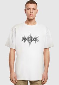 Hvid bomulds t-shirt med et fremtrædende sort pigget grafisk design og teksten "ANTIDOE." Korte ærmer og afslappet pasform.