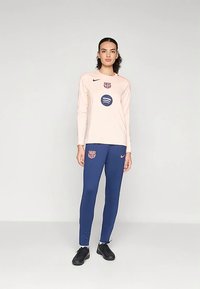 Ροζ μακρυμάνικη αθλητική μπλούζα με μπλε λεπτομέρειες, που φέρει λογότυπο και χορηγούς. Συνδυάζεται με μπλε παντελόνι και μαύρα ποδοσφαιρικά παπούτσια.