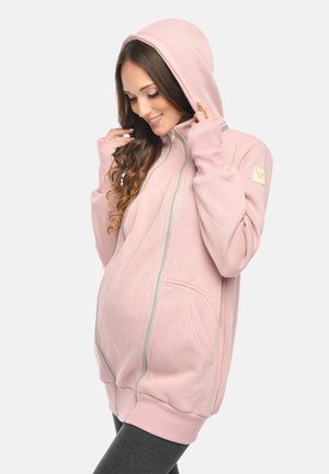 Pregnant woman in a pink maternity hoodie with front zipper pockets and thumbholes, adjusting the hood, smiling and looking down.
