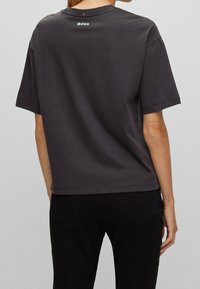 Osoba v volném tmavě šedém tričku s nápisem "BOSS" vytištěným na horní části zad, doplněném černými kalhotami, viděná zezadu.