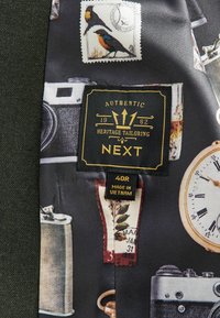 Crni materijal s vintage ilustracijama, uključujući kamere, markice i satove. Oznake upućuju na marku "NEXT", veličinu "40R" i podrijetlo "Vijetnam."