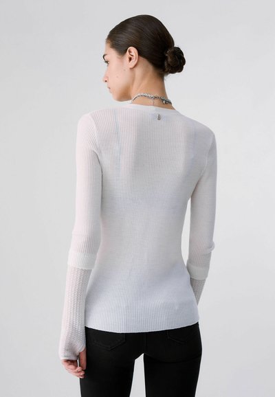 Femme aux cheveux foncés en chignon, portant un t-shirt blanc à manches longues côtelé et un pantalon noir, vue de dos sur un fond clair uni.
