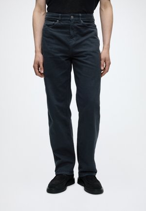 Personne portant un jean noir droit, des chaussures noires et une chemise noire rentrée, debout devant un fond blanc uni.