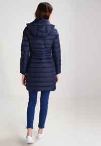 Middengroene pufferjas met een capuchon, voorzien van een gewatteerde structuur en lange mouwen, gecombineerd met blauwe jeans en witte sneakers.