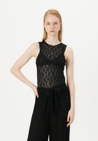 Svart blonde bodysuit med høy krage og ermeløs design, med et blomster mønster. Kombinert med høyt liv sorte vide bukser.
