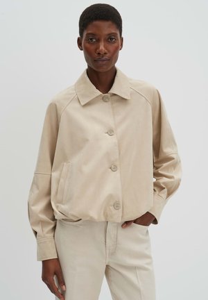 Donna con capelli corti che indossa una giacca beige con bottoni e pantaloni abbinati, in piedi con una mano in tasca davanti a uno sfondo semplice.