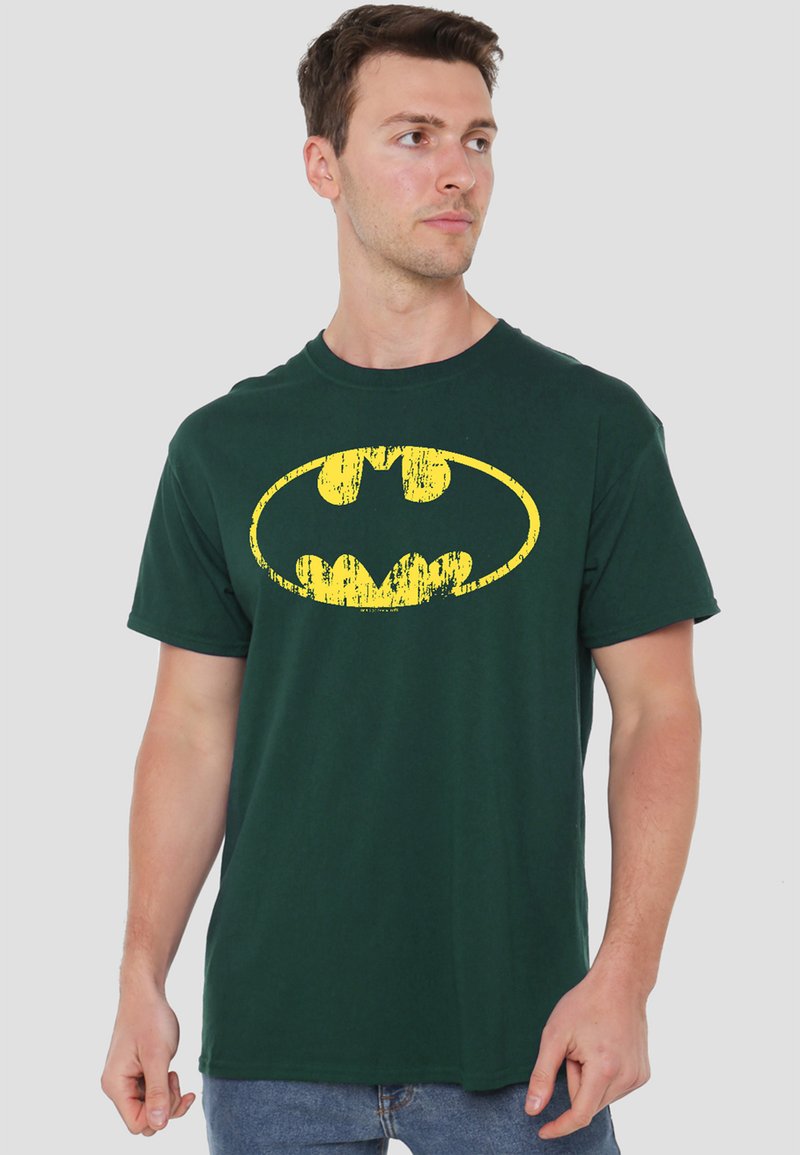 Young man wearing dark green t-shirt with a distressed yellow Batman logo, standing against a plain light background, looking right.