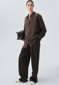 Camicia marrone con colletto, abbinata a pantaloni larghi marroni. Il modello tiene una pochette nera e indossa scarpe chiare.