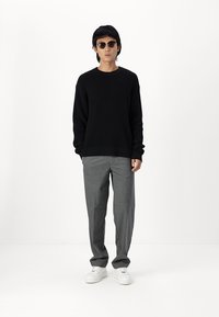 Fekete kötött pulóver, szürke nadrág és fehér sneaker. A modellen kerek napszemüveg van. A háttér sima fehér.