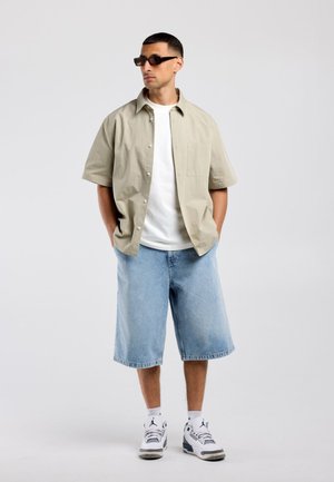 Man met zwarte zonnebril, beige korte mouw blouse met knopen over wit T-shirt, lichtblauwe denim shorts, witte sokken en witte sneakers.