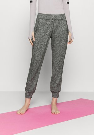 Personne portant un pantalon de jogging gris et un haut à manches longues clair, debout pieds nus sur un tapis de yoga rose sur fond blanc.