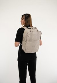 Beige ryggsäck med två remmar, ett handtag på toppen, framficka och logopatch. Slät textur mot en neutral bakgrund.