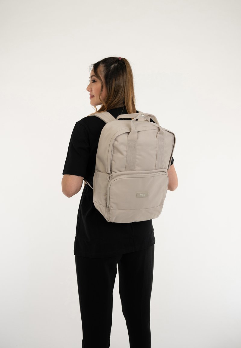 Beige ryggsäck med två remmar, ett handtag på toppen, framficka och logopatch. Slät textur mot en neutral bakgrund.