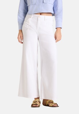 Femme portant un pantalon blanc taille haute à jambes larges, des sandales tressées dorées et une chemise courte bleu clair à manches longues.