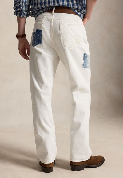 Άνδρας που φορά λευκό τζιν με μπλε denim επιδιορθώσεις στις πίσω τσέπες, καφέ ζώνη, καφέ μποτάκια και μπλε καρό πουκάμισο σε απλό φόντο.