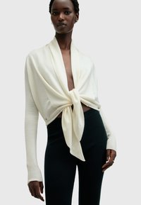 Suéter blanco con diseño anudado al frente, mangas largas con textura acanalada, combinado con pantalones negros ajustados. Tela lisa, estilo minimalista.