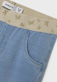 Light blue denim shorts with a beige waistband, featuring gold butterfly patterns. Includes a front pocket and subtle stitching details.