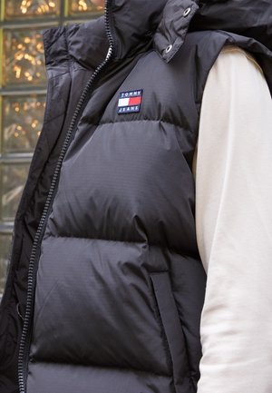 Gros plan d’une personne portant un gilet matelassé noir Tommy Jeans sur une chemise légère à manches longues, affichant le logo de la marque sur la poitrine.