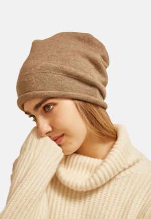 Berretto di maglia marrone con un design slouchy. Presenta una texture liscia e un tessuto morbido; abbinato a un maglione a coste di colore crema.