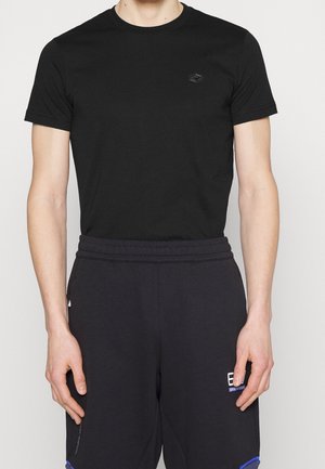 Man wearing plain black short-sleeve crew neck t-shirt and black sweatpants with small white text logo on left thigh.