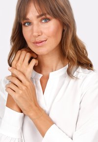 Vit blus med krage och långa ärmar. Guldarmband med en mycket fin kedjedesign på handleden. Slät hy och naturlig sminkning.