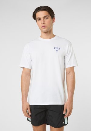 Jonge man staand, draagt een wit FILA T-shirt met korte mouwen en zwarte korte broek, neutrale uitdrukking, kleine kruis-tatoeage op de linker onderarm.