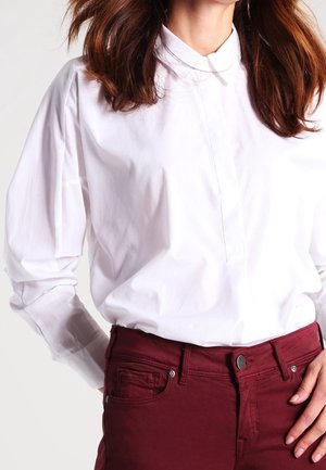Femme portant une chemise blanche à manches longues, rentrée dans un pantalon rouge foncé taille haute, les mains reposant le long du corps.
