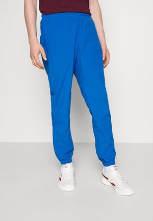 Person iført lyseblå joggingbukser, hvide sneakers med røde og sorte accenter, og en mørkerød skjorte mod en ensartet lys baggrund.