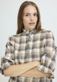 Kariertes Flanellhemd in Beige-, Grau- und Brauntönen. Es verfügt über einen rüschierten Kragen und eine Knopfleiste, mit hochgerollten Ärmeln und einem Stoff-Texture.