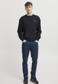 Črna trenirka z žepom na sprednji strani in teksturirano tkanino, v kombinaciji z mornarsko modrimi slim-fit hlačami. Model nosi temne športne čevlje.