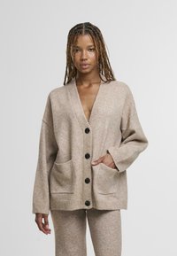 Beżowy sweter na cardigan z dekoltem w serek, czarnymi guzikami i dwoma przednimi kieszeniami. Miękka tekstura, luźny krój i prążkowane wykończenia.