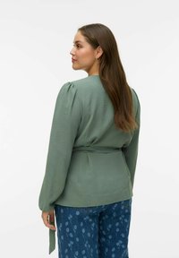 Grønn bluse med puffede ermer og knytebelte i midjen, kombinert med blå jeans med blomsterprint. Jevn tekstur og avslappet passform.
