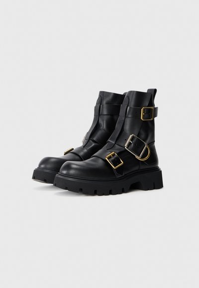 Botas de cuero negro hasta el tobillo con tres hebillas, suelas rugosas y acabados suaves, con acentos de hardware dorado.