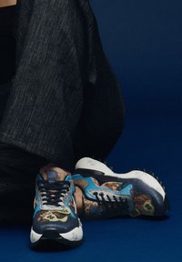 Zapatillas multicolores con un upper de tela texturizada, que presentan tonos de azul y marrón, patrones intrincados y suelas de goma negra con pinchos.